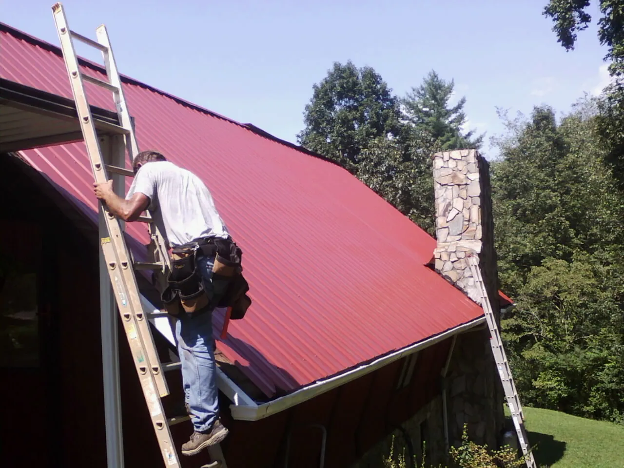 Roofer on a ladder beside a red metal roof and stone chimney — Soffit & Fascia Repair by Roofing Brothers in Rockvale